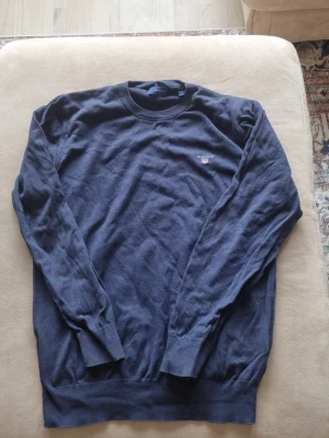 Mörkblå stickad tröja från GANT - Snygg mörkblå stickad tröja från GANT med rund halsringning och broderad logga på bröstet. Tröjan har långa ärmar och ribbade muddar vid ärmslut och nederkant. Perfekt för dig som gillar klassisk och stilren design.