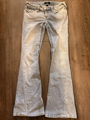 Ljusblå bootcut jeans, strl 25, från True Religion - Säljer ett par ljusblå bootcut jeans från True Religion med klassiska femficksdetaljer och snygga knappar på bakfickorna. Jeansen har en utsvängd passform och är tillverkade i denim med coola sömmar och patch på bakfickan. Knappt använda. Fräscha nu på våren!