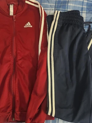 Adidas loungewear set röd/blå - Adidas loungewear set med röd zip-jacka och blå byxor. Jackan har vita ränder längs ärmarna och Adidas-logga på bröstet. Byxorna är mörkblå med vita ränder längs sidorna och resår i midjan. Perfekt för chill dagar hemma.