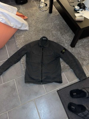 Mörkgrön overshirt från Stone Island - Snygg mörkgrön overshirt från Stone Island med dragkedja framtill och två bröstfickor. Klassisk krage och ikonisk logotyp-patch på vänster ärm. Tillverkad i ett slitstarkt material som ger en clean och stilren look. Lite solblekt på ryggen men inget man tänker på.