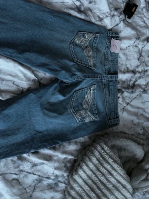 Blå bootcut jeans med broderade fickor - Snygga blå bootcut jeans från Miss Me med slitningar på benen och broderade detaljer på bakfickorna. Jeansen har klassisk femficksdesign och är tillverkade i denim med stretch för extra komfort. Perfekta för dig som gillar en trendig och avslappnad stil.
