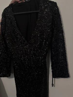 Svart glittrig klänning med v-ringning - Snygg svart klänning med glittriga paljetter och djup v-ringning. Klänningen har trekvartsärm och figurnära passform, perfekt för att sticka ut. Materialet är syntet och paljetter som ger en lyxig känsla.