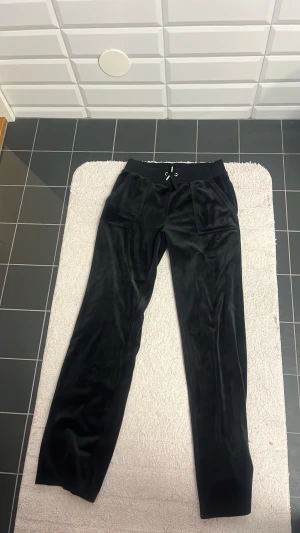 Svarta mjukisbyxor i velour storlek:S  - Snygga svarta mjukisbyxor i mjukt material med broderad logga på sidan. Byxorna har en bekväm passform och dragsko i midjan för justerbarhet. Aldrig använda och jag köpte mjukisbyxorna från Nelly och jag köpte de för 1200 kronor men kan tänka mig att sälja för 600 kronor. 