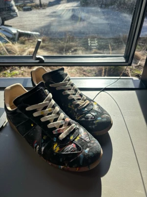 Svarta sneakers med färgstänk - Maison Margiela sneakers i svart läder med beige snörning och gummisula. Skorna har ett unikt färgstänksmönster i gult, blått, vitt och rött över hela ovandelen. Klassisk låg modell med retrovibe och konstnärlig touch. Jag har haft de ungefär 4 veckor men har tröttnat på de och vill helst sälja dom så jag kan köpa några andra. Vill helst inte sänka priset så mycketköpte för 2900kr begagnade.