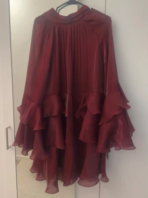 Klänning - Långärmad klänning med volanger nedtill och på ärmarna. Klänningen är vinröd och har hög krage. Den har oversize fit, glansigt material och kort längd. Går att knyta baktill. Strl 38  Använd en gång endast