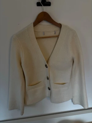 Beige kofta med knappar och fickor - Snygg beige kofta med tre stora knappar framtill och två praktiska fickor. Koftan har ribbade muddar och en djup v-ringning. Perfekt att slänga över en t-shirt för en chill look.