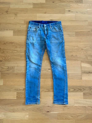  jeans från Levi's - Säljer ett par blå jeans från Levi's med slitna detaljer och klassisk femficksmodell. Jeansen har en smal passform och tydliga slitningar framtill för en trendig look. Perfekta för dig som gillar en avslappnad och cool stil.