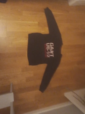 Svart GANT hoodie med tryck - Svart hoodie från GANT med stort vitt och rött tryck på bröstet där det står 'GANT USA 1949'. Klassisk modell med långa ärmar och bekväm passform. Perfekt för en avslappnad och sportig stil.
