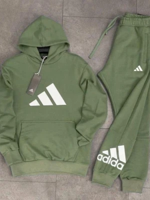 Grön Adidas loungewear set - Snyggt loungewear set från Adidas i olivgrönt med matchande hoodie och byxor. Båda plaggen har stora vita Adidas-loggor framtill. Hoodien har känguruficka och byxorna har dragsko i midjan för skön passform. Perfekt för chill dagar hemma.