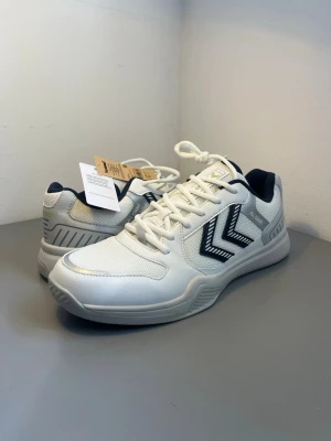 Vita Hummel sneakers med svarta detaljer - Snygga vita sneakers från Hummel med svarta och grå detaljer samt märkets klassiska pilar på sidan. Skorna har en låg profil, mesh- och syntetmaterial, samt vadderad häl och plös för extra komfort. Perfekta för en sportig och stilren look.