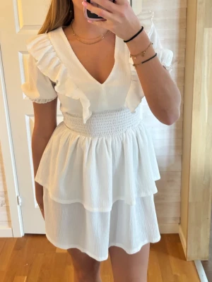 Vit volangklänning med v-ringning - Superfin vit kortklänning med v-ringning, volangdetaljer på axlar och bröst samt puffärmar. Klänningen har smockad midja och dubbla lager i kjolen för extra volym. Perfekt för sommarens alla tillfällen. 