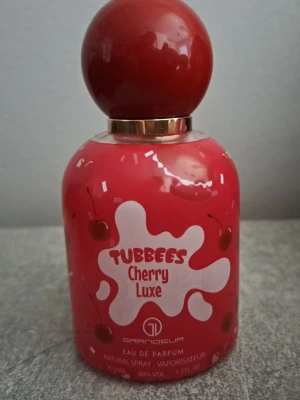 Tubbees Cherry Luxe parfym - Tubbees Cherry Luxe är en parfym med söt körsbärsdoft. Flaskan är klarröd med ett stort, runt lock och detaljer i guld. Designen har körsbärsmotiv och en lekfull vit logga på framsidan. Perfekt för dig som gillar fruktiga och färgstarka dofter.