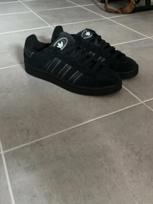 Svarta Adidas Campus sneakers - Säljer ett par svarta Adidas Campus sneakers i mocka med klassiska vita ränder på sidorna och svart sula. Skorna har låga skaft och svart snörning. Adidas-logga på plösen och hälen. Perfekta för en clean och stilren look. Helt nya typ