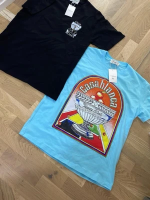 Casa t-shirts med tryck - Två snygga t-shirts från Casablanca. En svart med stort grafiskt tryck av tennisracketar och texten 'Casa Blanca', samt en ljusblå med färgstarkt motiv och 'Casablanca Tennis Club' på bröstet. Båda har rund hals och korta ärmar. Perfekta för dig som gillar streetwear och sportig stil.
