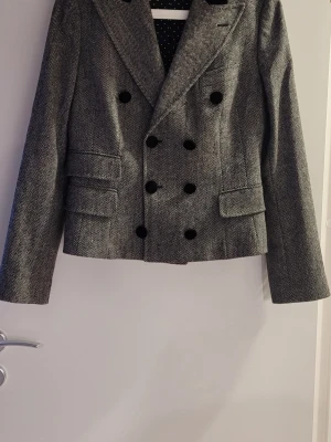 Grå dubbelknäppt tweedkavaj i Ull från D&G - Snygg grå kavaj med fiskbensmönster och dubbelknäppning. Stors svarta knappar framtill och på ärmarna, samt två fickor med lock. Klassisk krage och figurnära passform. Perfekt för en stilren look. IT storlek 44 som motsvarar en EU 38. 89% Ull, som inte sticks.