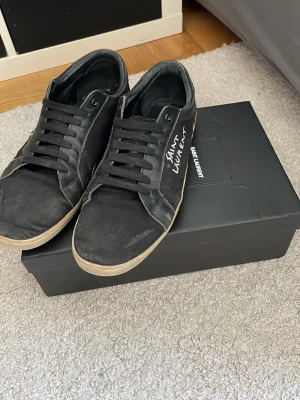 Svarta Saint Laurent sneakers - Svarta låga sneakers från Saint Laurent med vit logga på sidan. Skorna har svart snörning, gummisula i beige och är tillverkade i läder. Klassisk och stilren design som passar till det mesta. Perfekta för dig som gillar exklusiva streetwear-vibbar.