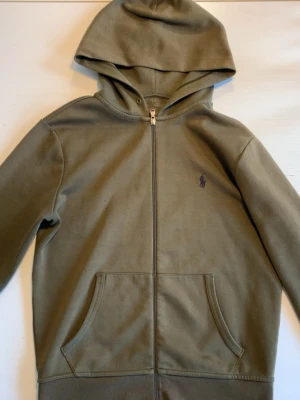 Olivgrön hoodie från Polo Ralph Lauren - Snygg olivgrön hoodie med dragkedja från Polo Ralph Lauren. Klassisk design med huva, kängurufickor och den ikoniska broderade loggan på bröstet. Tillverkad i mjuk bomullsblandning som är skön att ha på sig. Perfekt för en avslappnad och stilren look. Tröjan har en liten missfärgning men man märker det verkligen inte när den används. Tveka inte på att fråga!🤗OBS pris kan diskuteras!