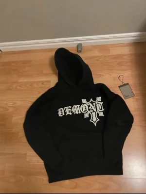 Svart hoodie med DEMONT-tryck - Slut såld på deras hemsida, Snygg svart hoodie från Demont med stort vitt DEMONT-tryck och korsdetalj på bröstet. Klassisk passform med huva och känguruficka framtill. Perfekt för dig som gillar streetwear och vill sticka ut med en unik design.