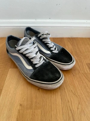 Vans Old Skool svart och grå sneaker - Klassiska Vans Old Skool sneakers i svart och grått med vit sidosnöre och vit sula. Ovandelen är i mocka och canvas, med vita skosnören och den ikoniska vågformen på sidan. Perfekta för en streetwear-look.