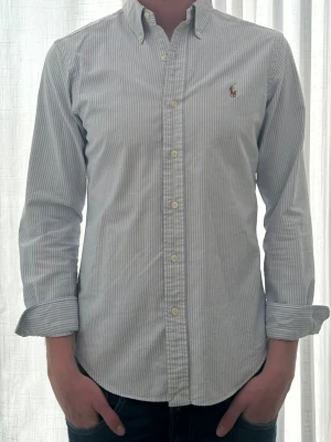 Randig skjorta från Ralph Lauren - Klassisk ljusblå och vit randig skjorta från Ralph Lauren med button-down krage och broderad logga på bröstet. Skjortan har långa ärmar och knäppning framtill. Perfekt för en stilren look.