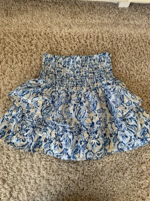 Blåvit volangkjol med mönster - Supersöt kort kjol med blått och vitt mönster och bred smock i midjan. Kjolen har volanger som ger en flowy känsla och är perfekt för varma dagar. Tillverkad i ett lätt och mjukt material som känns skönt mot huden.