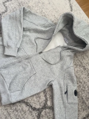 Grå hoodie från C.P. Company - Snygg grå hoodie från C.P. Company med dragkedja, huva med dragsko och två stora fickor framtill. Ärmen har en ikonisk svart linsdetalj och en extra ficka med dragkedja. Tillverkad i mjuk bomullsblandning för maximal komfort och perfekt för sommaren. Den är helt oanvänd. Den kan möjligtvis också passa vissa som har XS.