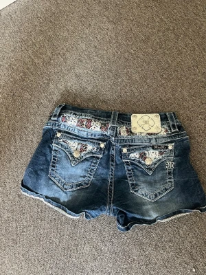 Blå jeansshorts från Miss Me - Säljer ett par blå jeansshorts från Miss Me med snygga broderade detaljer och dekorativa knappar på bakfickorna. Shortsen har midwaist passform och coola slitningar för en avslappnad look. Perfekta till sommaren!