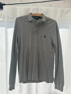 Grå långärmad skjorta/ tröja från Polo Ralph Lauren - Snygg grå långärmad skjorta/ tröja från Polo Ralph Lauren med klassisk krage och knappar framtill. Skjortan har broderad logga på bröstet och är tillverkad i mjuk bomull. Perfekt för en stilren och avslappnad look.