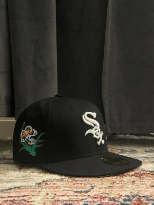 Svart White Sox keps med broderade fjärilar - Säljer en svart snapback keps från New Era med White Sox-logga framtill och broderade fjärilar i färger som gult, orange och vitt på sidan. Kepsen har platt skärm och klassisk passform. Perfekt för dig som vill sticka ut med coola detaljer.