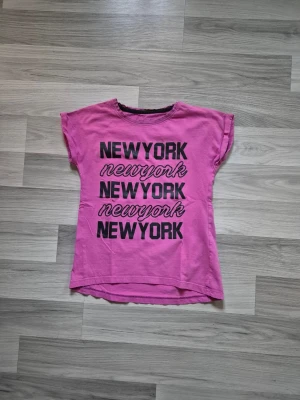 Rosa t-shirt med New York-tryck - Cool rosa t-shirt med svart New York-tryck i olika stilar på framsidan. T-shirten har korta ärmar och en avslappnad passform. Perfekt för dig som gillar streetstyle och vill sticka ut lite extra.