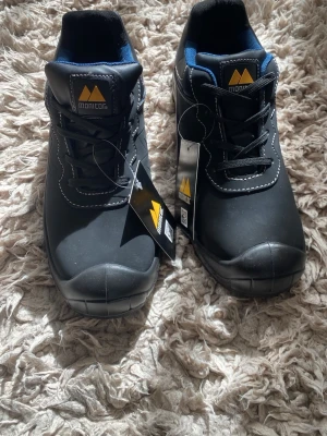 Svarta skyddsskor från Monitor - Svarta skyddsskor från Monitor med vita kontrastsömmar och blå insida. Skorna har en låg profil, rund tå och snörning framtill. Tillverkade i slitstarkt material med robust sula, perfekta för en aktiv vardag.