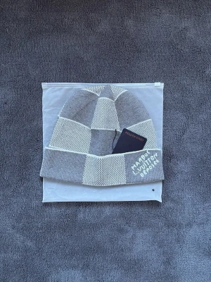 Marque L.Vuitton depcsee  - Stilren stickad mössa i grå/vit design från Louis Vuitton. Klassiskt mönster med diskret logga framtill.  Mjuk och varm – perfekt för kallare dagar. Skick: Nytt skick!