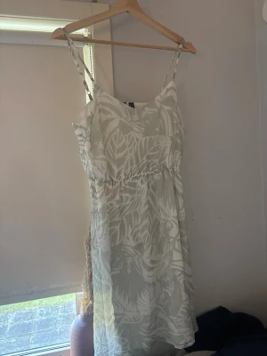 Vit sommarklänning med bladmönster - Luftig vit kortklänning med tunna axelband och ett snyggt bladigt mönster. Klänningen har en lätt v-ringning och resår i midjan för en bekväm passform. Perfekt för varma dagar och har en fräsch, ljus känsla.