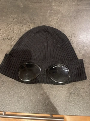 Svart C.P. Company goggle mössa - Säljer en svart ribbstickad mössa från C.P. Company med ikoniska goggle-linser på uppviket. Mössan är tillverkad i mjukt material och har en cool, urban look som sticker ut. Perfekt för dig som vill ha något unikt och trendigt.