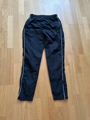 Svarta träningsbyxor med vita ränder - Svarta träningsbyxor med vita ränder längs sidorna och resår i midjan. Byxorna har en sportig look och är tillverkade i ett lätt och smidigt material, perfekt för träning eller fritid. Klassisk passform med elastiska benslut.