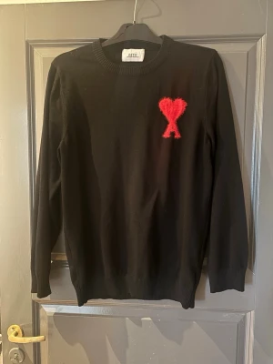 Svart stickad tröja från Ami Paris - Svart stickad tröja från Ami Paris med ett rött, fluffigt hjärta och bokstaven A broderat på bröstet. Tröjan har rund halsringning, långa ärmar och ribbade muddar. Perfekt för dig som gillar stilrena plagg med en twist. Fint skick utan defekter alls. Tryck köp nu