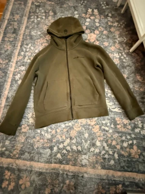 Olivgrön hoodie från Sail Racing - Säljer en olivgrön hoodie från Sail Racing med dragkedja framtill och två stora fickor. Hoodien har en broderad logga på bröstet och är tillverkad i mjukt bomullsmaterial. Perfekt för en avslappnad och sportig stil.