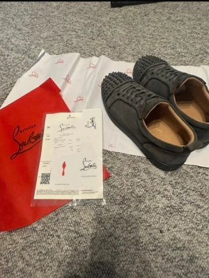 Gråa Christian Louboutin sneakers - Ett par diskreta grå klassiker med subtila metallinslag som fångar ljuset i rörelse. En av detaljerna har försvunnit och lämnar ett nästan omärkligt avtryck, medan bakpartiet bär spår av en liten förändring i materialet. Helhetsintrycket förblir stilrent och genomtänkt, där små egenheter smälter in och ger karaktär utan att ta över.
