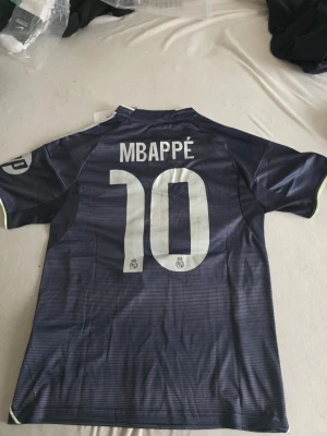 Real Madrid matchtröja Mbappé #10 - Säljer en mörkblå Real Madrid matchtröja från Adidas med Mbappé och nummer 10 på ryggen. Tröjan har vita detaljer, klubbmärke och sponsortryck på framsidan samt patchar på ärmarna. Materialet är lätt och andas, perfekt för fotboll eller träning.