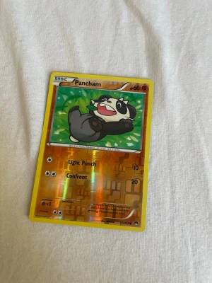 Pokémonkort Pancham holo - Säljer ett holografiskt Pokémonkort med Pancham-motiv. Kortet har gul ram och glittrande effekt, med attackerna Light Punch och Confront. Perfekt för samlare eller dig som vill utöka din Pokémonkort-lek. Kortet är officiellt från Pokémon TCG.