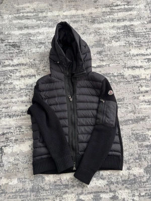 Svart pufferjacka från Moncler - Svart pufferjacka från Moncler med huva och dragkedja framtill. Jackan har stickade ärmar och muddar samt Moncler-logga på ena ärmen. Kombinerar quiltat material på kroppen med stickat tyg på ärmarna för en cool kontrast.