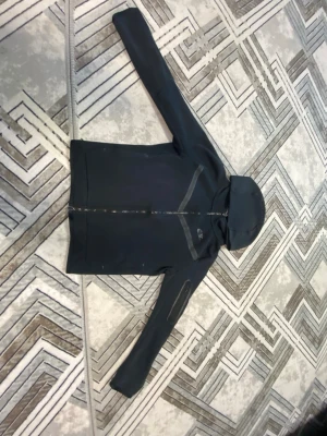 Svart Nike hoodie med dragkedja - Snygg svart hoodie från Nike med hel dragkedja och huva. Har en diskret Nike-logga på bröstet och en grå detalj över bröstet. Perfekt för en sportig och avslappnad stil.