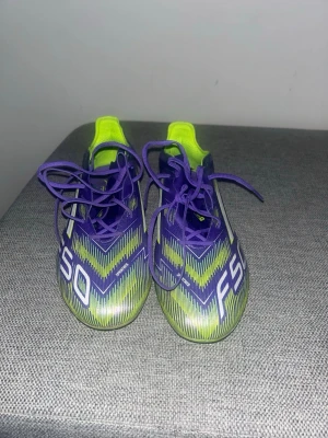 Adidas F50 lila fotbollsskor (konstgräs) - Säljer ett par Adidas F50 fotbollsskor i lila och neongult med vita detaljer Storlek 44. Skorna har snörning, låg profil och en sportig design med tydliga F50- och Adidas-loggor. Ovandelen är i syntetmaterial för bra bollkontroll och lätt känsla på planen. Har används endast 1 gång och är i perfekt skick. (Kommer med extra sulor & väska)