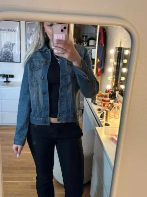 Klassisk blå jeansjacka - Säljer en klassisk blå jeansjacka med raka sömmar och bröstfickor med lock. Jackan har knappar framtill och vid ärmsluten, samt en figurnära passform som ger en snygg siluett. Perfekt att slänga över en t-shirt eller hoodie för en cool look.