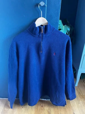 Marinblå halvzip från Ralph Lauren - Mörkblå stickad tröja från Ralph Lauren med halv dragkedja och hög krage. Liten röd broderad logga på bröstet. Långärmad och i mjukt material, perfekt för en stilig sommar outfit!