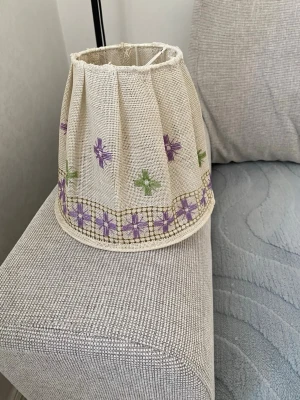 Beige lampskärm med broderi - En beige lampskärm i tyg med broderade mönster i lila och grönt. Skärmen har veckad form och dekorativa korsstygn längs nederkanten. Passar till mindre lampfötter och ger ett personligt intryck till rummet.
