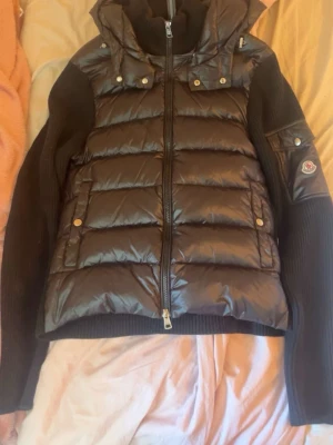 Svart dunjacka från Moncler - Snygg svart dunjacka från Moncler med stickade ärmar och huva. Jackan har quiltad front i glansigt material, dragkedja framtill och fickor med tryckknappar. Moncler-logga på ärmen och ribbstickade detaljer för extra stilpoäng.