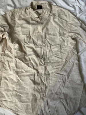 Beige linneskjorta från H&M - Stilren beige skjorta i linne från H&M med klassisk krage och knäppning framtill. Skjortan har lång ärm och en avslappnad passform, perfekt för en trendig och bekväm look. Den är inte använd, så det är därför den är kryngklig