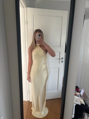 Ljusgul långklänning med halterneck - Elegant ljusgul långklänning med halterneck och figurnära passform. Klänningen har en slät yta med lätt glans och är ärmlös, perfekt för speciella tillfällen. Materialet känns svalt och följsamt mot kroppen.