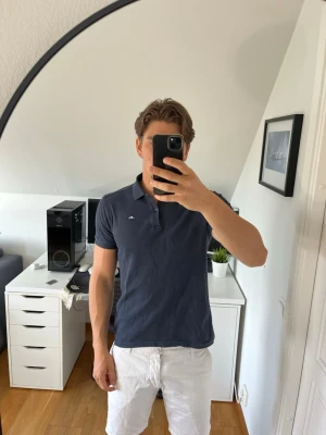 Mörkblå pikétröja från J.Lindeberg - Snygg mörkblå pikétröja från J.Lindeberg med klassisk krage och knappar framtill. Tröjan har korta ärmar och en liten vit logga på bröstet. Hör gärna av dig vid frågor!